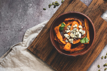 Baked pumpkin slices with blue cheese and herbs in a ceramic bowl on a wooden cutting board. Rural scene. Top view, flat lay. Toned photo