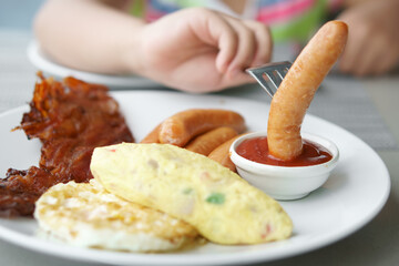 hand of child or kid holding fork dipping sausage with ketchup or tomato sauce and fried egg with omelet and grilled bacon on white dish for morning food eating or breakfast at home or restaurant