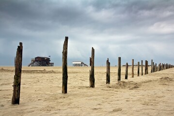 Two stilt houses in the background and stockades in line on sandy beach with traces of the North sea in Sankt Peter-Ording, Germany.