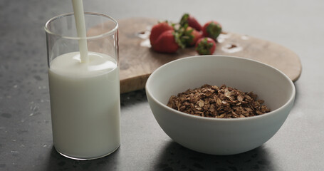 pour yogurt into glass next to white bowl with chocolate granola, strawberries on a concrete countertop