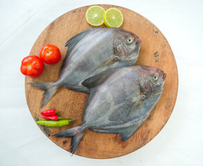 side view of black pomfret fish dish cooking with various ingredients. Fresh raw fish decorated with lemon slices and tomato on a wooden pad,white background.