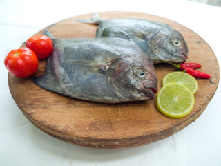 side view of black pomfret fish dish cooking with various ingredients. Fresh raw fish decorated with lemon slices and tomato on a wooden pad,white background.