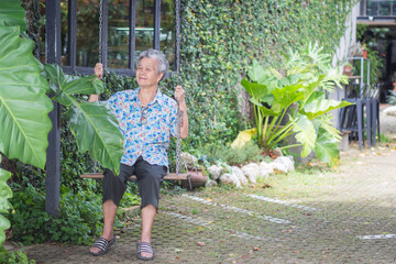 An elderly Asian woman smiling and look at camera while sitting on a swing in a garden. Space for...
