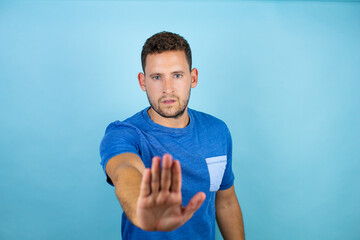 Young handsome man wearing blue casual t-shirt over isolated blue background serious and doing stop sing with palm of the hand.