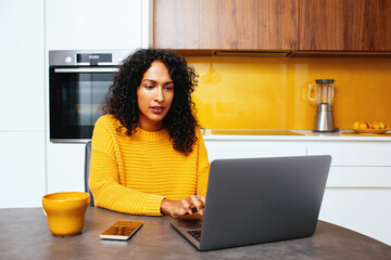 Portrait of a mid woman with black curly hair usingt laptop computer in kitchen