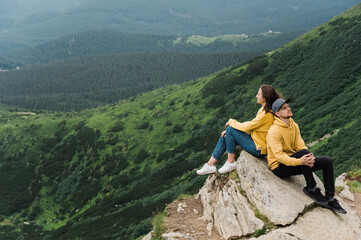 Naklejka premium Inspirational landscape. Love in the mountains. Happy couple in love sits on the edge of the cliff back to back on the big stone with an amazing mountains view on the background