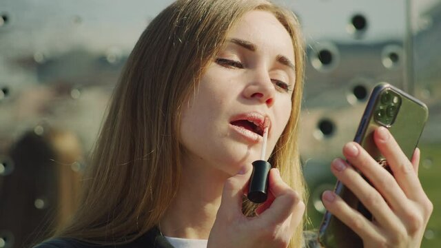 Young woman applying lipstick in front of a phone at the street. Red Helium with Carl Zeiss CP.2.