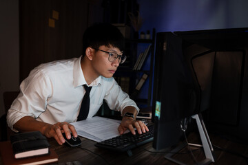 Young pensive asian man working late concentrated and serious in front of computer at night in dark office, Late night working or studying concept