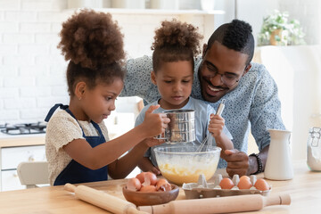 Happy young african american dad watching two adorable mixed race kids siblings preparing dough for homemade pastry, enjoying weekend family activity together in kitchen, culinary recipe concept.