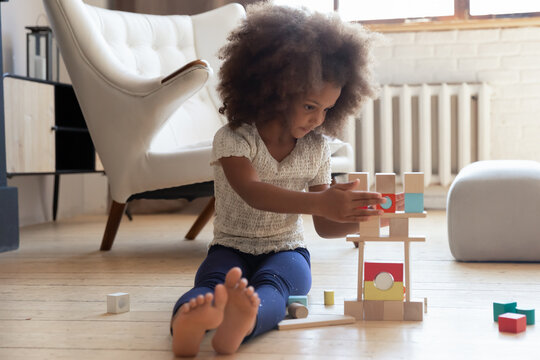 Full length cute small african ethnicity barefoot kid girl sitting on warm floor, building tower with wooden blocks in living room. Focused adorable little biracial child playing alone at home. - Powered by Adobe