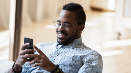 Relaxed 30s african american man in eyeglasses sitting on sofa, involved in online communication, using mobile application at home, smiling guy web surfing information or playing game on smartphone.