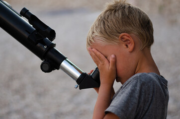 Young boy on a pebble beach looking through a telescope at the evening sky, covering one eye with his hand.