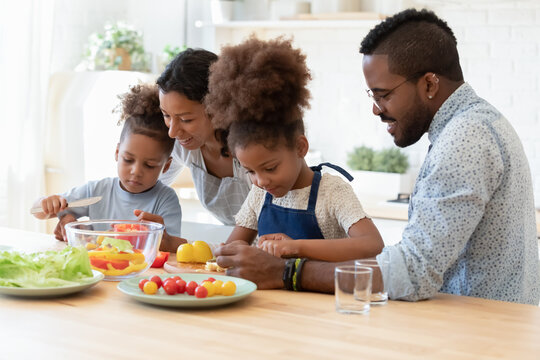 Happy Mixed Race Young Parents Teaching Little Kids Siblings Cooking Fresh Healthy Vegetarian Food At Home, Chopping Vegetables Together In Modern Kitchen, Positive Bonding Family Weekend Pastime.