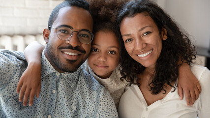 Head shot close up portrait of smiling cute african ethnicity little kid girl cuddling mixed race parents. Affectionate couple enjoying sweet tender moment with child daughter, posing for photo.