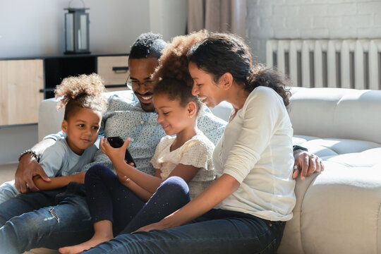 Addicted to technology happy bonding affectionate african american family couple watching funny video content on smartphone with joyful little kids siblings, leaning on couch in modern living room.