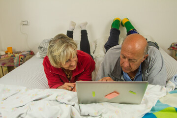 elderly husband and wife on the bed in their bedroom talking and laughing and having fun watching the laptop computer passing joyful moments