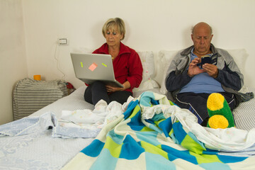 elderly husband and wife sitting on the bed looking at both the smartphone and the laptop with total indifference without worrying about each other
