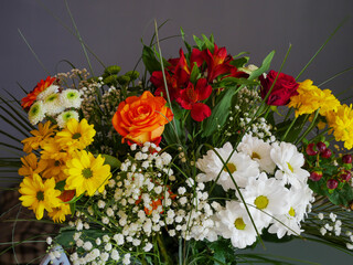 Joli bouquet de fleurs, jaunes, blanches oranges et rouges  accompagnées de gerbes vertes,  sur fond gris. 