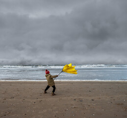 Young boy struggling with large yellow umbrella on beach in stormy weather. Winter beach scene.