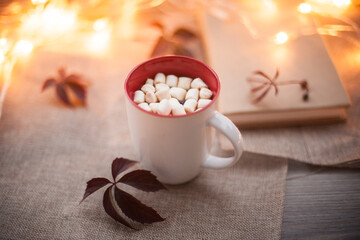 White glass with cocoa and marshmallows on a wooden table with autumn leaves and bokeh in the background. Photos in warm autumn colors.