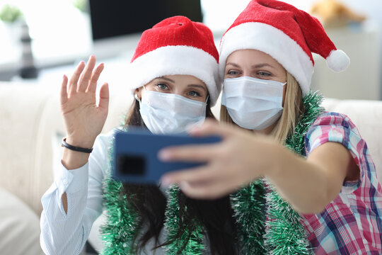 Two young women in santa hats and protective masks on faces are holding phone at home portrait. Online communication during covid 19 pandemic concept.