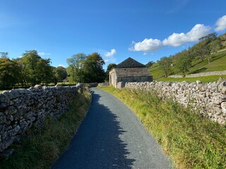 Country lane, with dry stone walls, grass verges, and farm buildings, leading to, Hawkswick, Skipton, UK