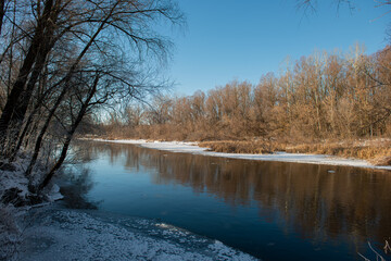 Naklejka na ściany i meble Silhouettes of trees of deciduous forest and river with stripes of snow and ice along the banks on a sunny day. .