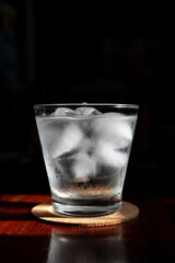 Glass of water with ice cubes on wooden table; shades of sunlight through from window.