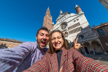 happy tourist couple visiting the city of Cremona taking selfie in the middle of main square of Cremona with Cathedral, baptistery and Torrazzo bell tower . 