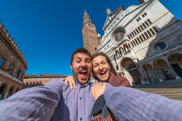 happy tourist couple visiting the city of Cremona taking selfie in the middle of main square of Cremona with Cathedral, baptistery and Torrazzo bell tower . 