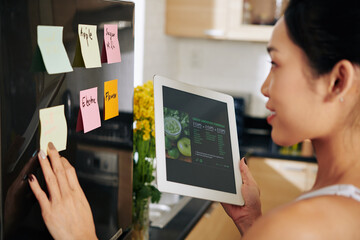 Young woman reading recipe on tablet computer and sticking notes on refrigerator before ordering...