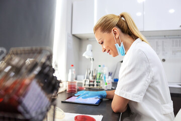 Innovative experienced female lab assistant in white sterile uniform with surgical mask and rubber gloves on sitting in laboratory and using tablet to enter test results.