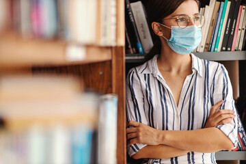 Young attractive freshman girl with face mask on standing in library with arms crossed and looking away. Corona virus pandemic concept.