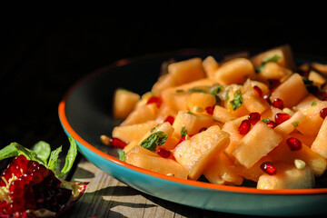 Sliced melon, poured with honey with pomegranate seeds and mint leaves, on a large ceramic plate, and a dark background.