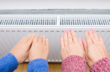 Woman and child wearing warm clothes and warming hands near heating radiator. Heating season at...
