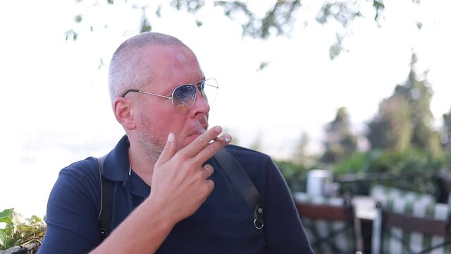 Video, an adult man in sunglasses smokes a cigarette in a cafe at a table, on a sunny day in summer