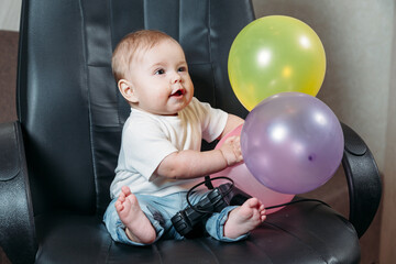 Small child sits on a chair with a joystick from a video game and holds balloons. The concept of playing with a small child.
