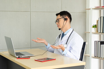 young asian doctor wearing headphone and using computer to give consult to the patient via video call in the hospital office. telemedine and healthcare concept