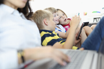 Three children sit side by side on couch and play on smartphones. Boy and girl look at phone screen in surprise. Woman is typing text on laptop next to children.