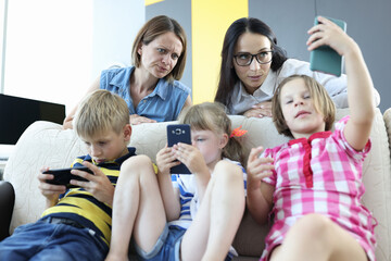 Three children sit side by side on couch and play games on phone. One woman look unhappily at daughter phone screen. Mother in glasses look surprised at girl phone.