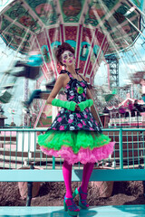 Girl with art make-up and hairstyle standing near the carousel. Happy young clown girl. Doll girl.