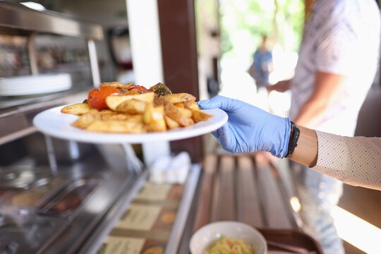 Female hand in blue medical glove hold white plate with fried potatoes and stewed vegetables. Self service restaurant with tray.