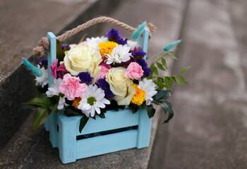 A bouquet of beautiful multi-colored flowers in a blue wooden box on the steps of the stairs. Present.
