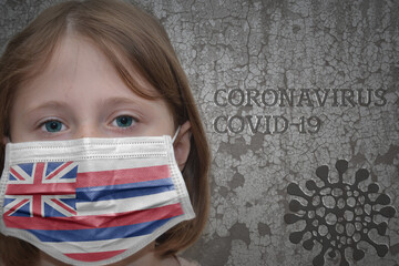 Little girl in medical mask with hawaii state flag stands near the old vintage wall with text coronavirus, covid, and virus picture. Stop virus concept