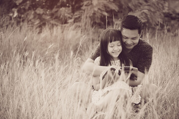 Kind asian father sitting on grass with little daughter sitting on him and reading a book.Vintage color style