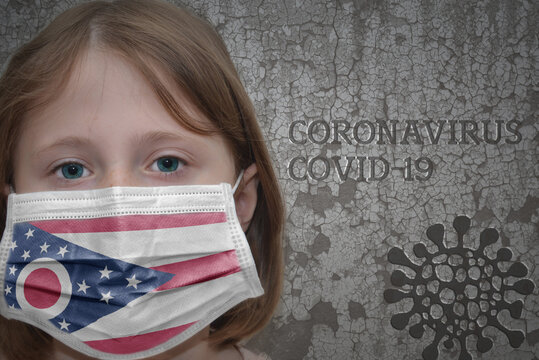 Little girl in medical mask with ohio state flag stands near the old vintage wall with text coronavirus, covid, and virus picture. Stop virus concept