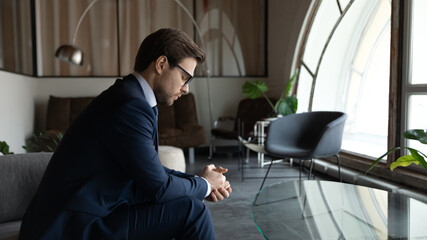 Side view nervous worrying 30s businessman in formal suit and eyewear sitting on couch with folded...