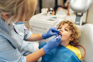 Cute young boy visiting dentist, having his teeth checked by female dentist in dental office.