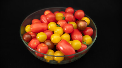 
Small cocktail tomatoes, multicolored, round and oval, in a transparent glass container, on black background, close-up	