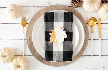 Autumn harvest or thanksgiving dinner table setting with plates, flatware, buffalo plaid napkin, pumpkins and decor. Top view on a white wood background.